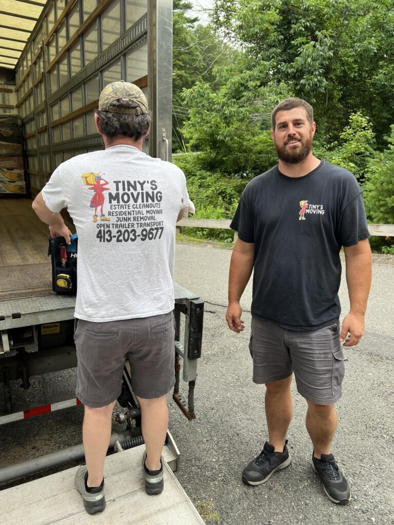 Two men standing next to moving truck one is facing the camera smiling the other has his back to the camera wearing a shirt that says Tiny's Moving